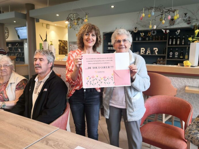 Photo d'un groupe de résidents d'une maison de retraite. Deux femmes au centre tiennent une affiche de remerciement pour une collecte caritative. La scène se déroule dans un espace commun décoré pour Pâques avec des œufs suspendus. L'ambiance est conviviale.